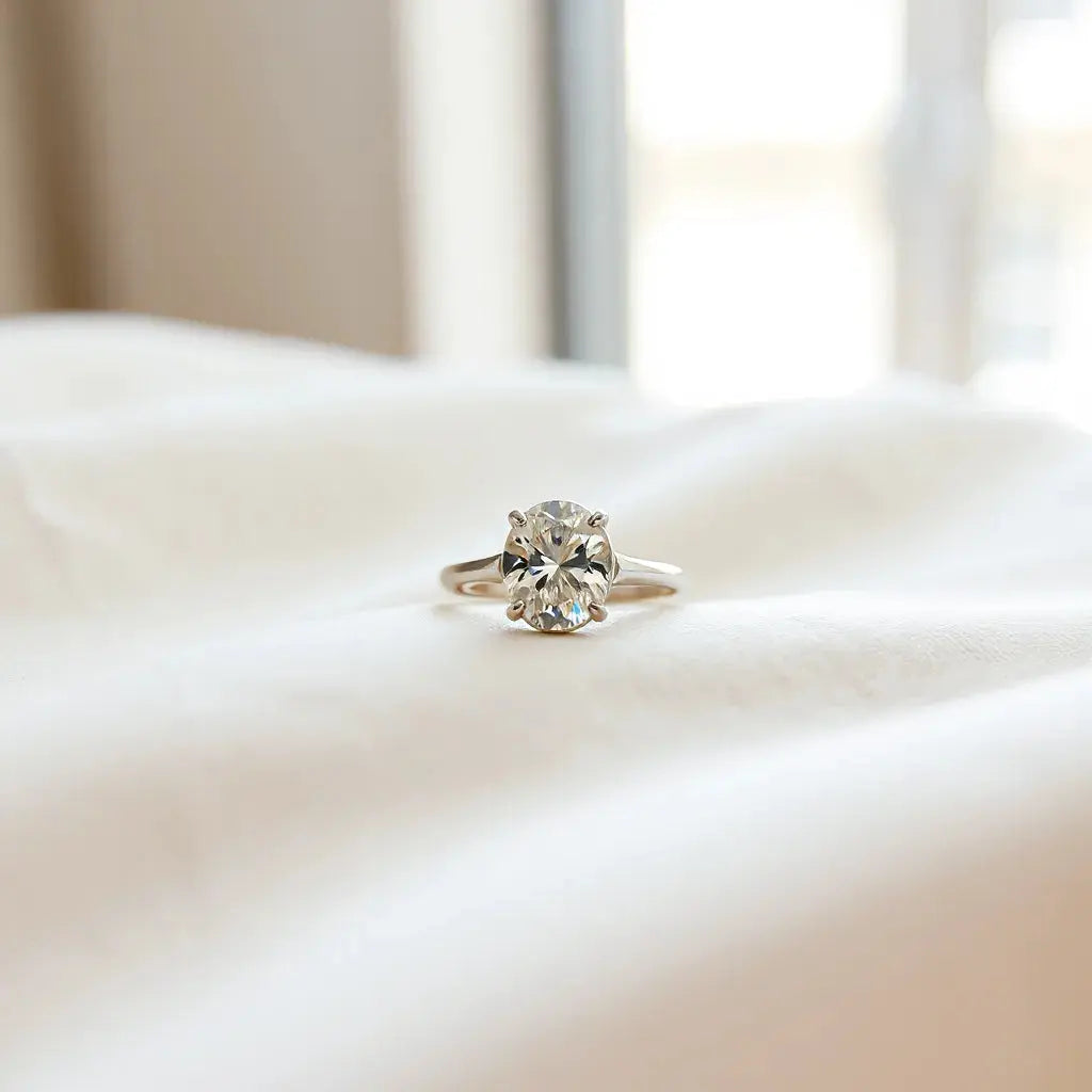 Oval diamond ring on neutral fabric with window light highlighting the oval sparkle and band details