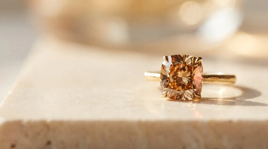 A champagne diamond solitaire ring resting on warm travertine stone, bathed in soft natural light that illuminates the diamond's rich golden and cognac tones.