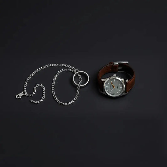 Minimal men’s jewellery set with silver chain, ring, bracelet, and watch on a dark surface