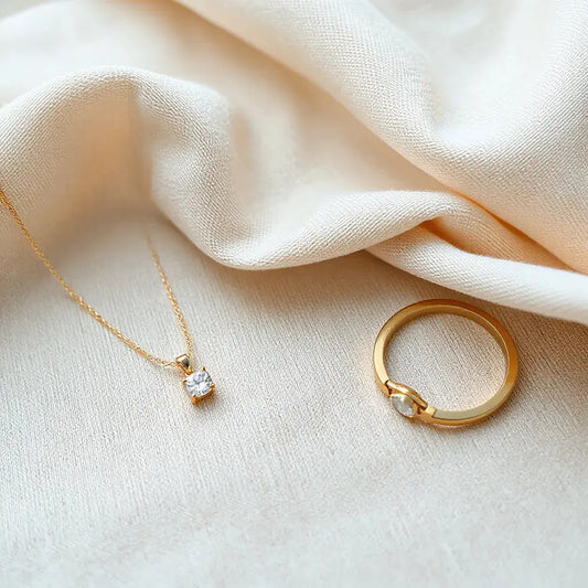 Gold pendant necklace, diamond solitaire ring, and fine bracelet on neutral fabric with soft studio light